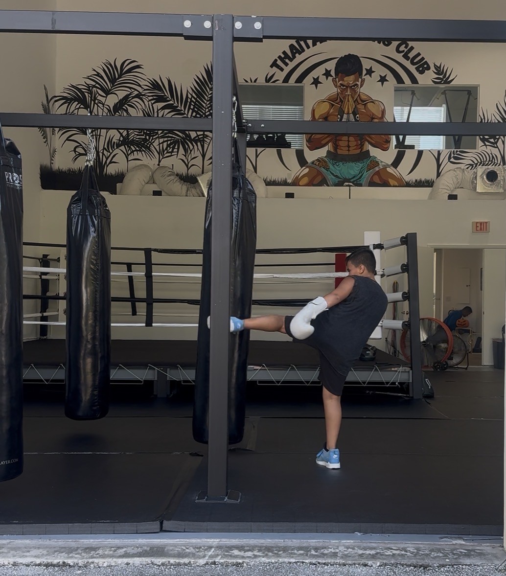 Student throwing a kick on the heavy bag with the ring behind him at Thaitan Boxing Club